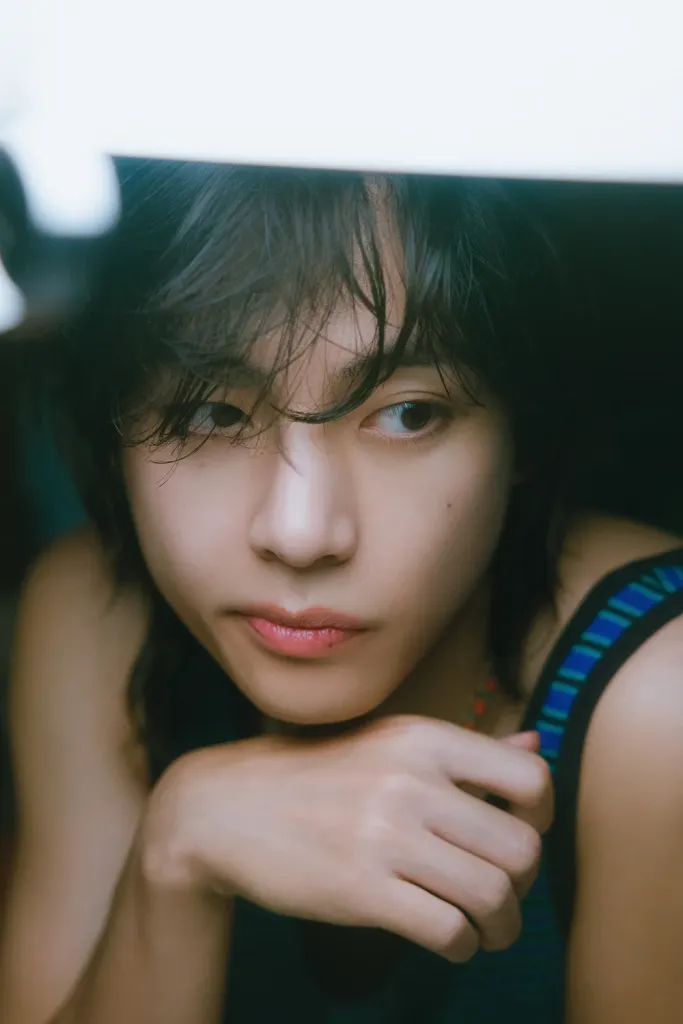 BTS V with soft features, slightly tousled hair, and a pensive expression, resting their chin on their hand.