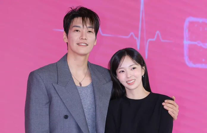 Two smiling individuals pose together at an event, with a pink background featuring a heartbeat line graphic.