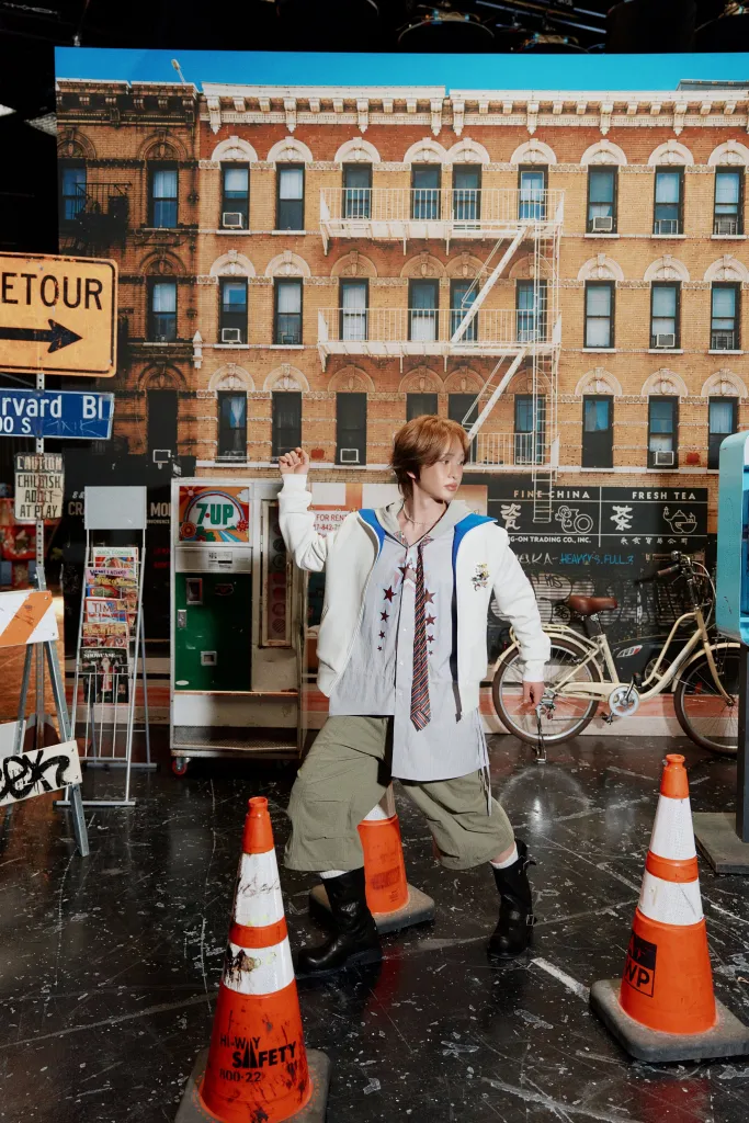 Onew posing in a stylish outfit with a white oversized jacket, tie, and baggy pants, standing among orange traffic cones in front of a cityscape backdrop featuring a brick building and storefronts.