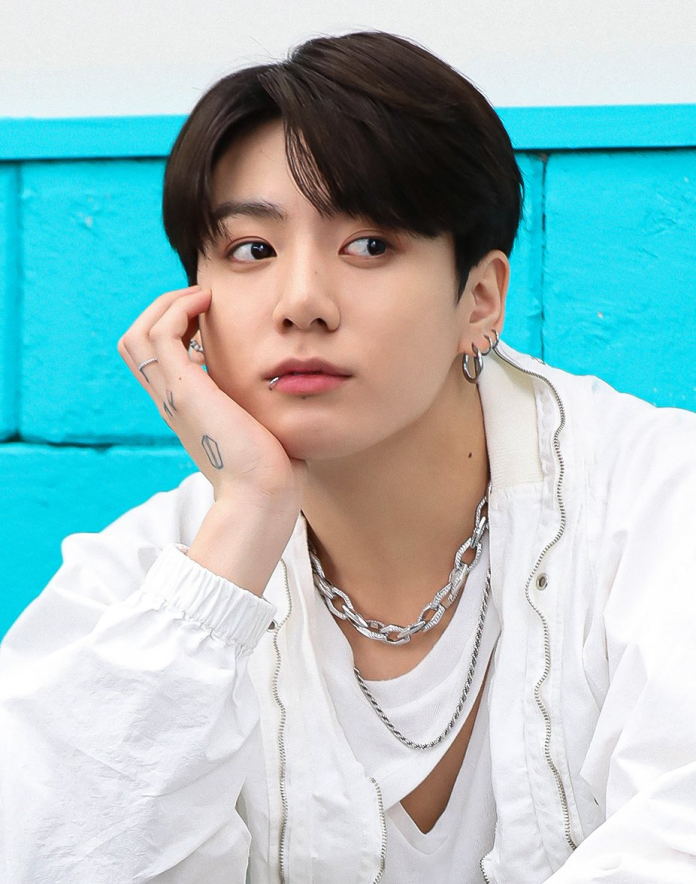 Jung Kook with dark hair wearing a white jacket and layered silver necklaces, resting their chin on their hand in front of a blue wall.