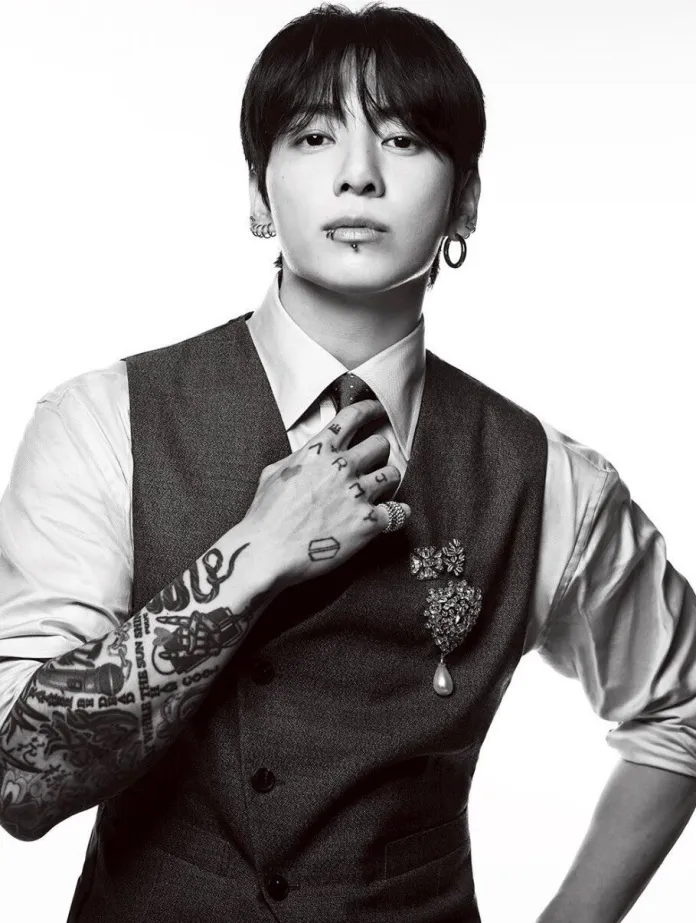 Jung Kook with long hair and earrings, wearing a vest over a collared shirt, showcasing tattoos on their arm and a decorative brooch on the vest.
