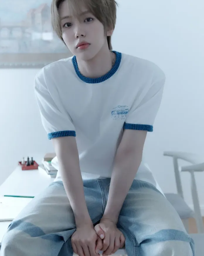 Cha Woongki sitting on a desk, wearing a white t-shirt with blue trim and faded jeans, looking thoughtfully at the camera in a softly lit room.