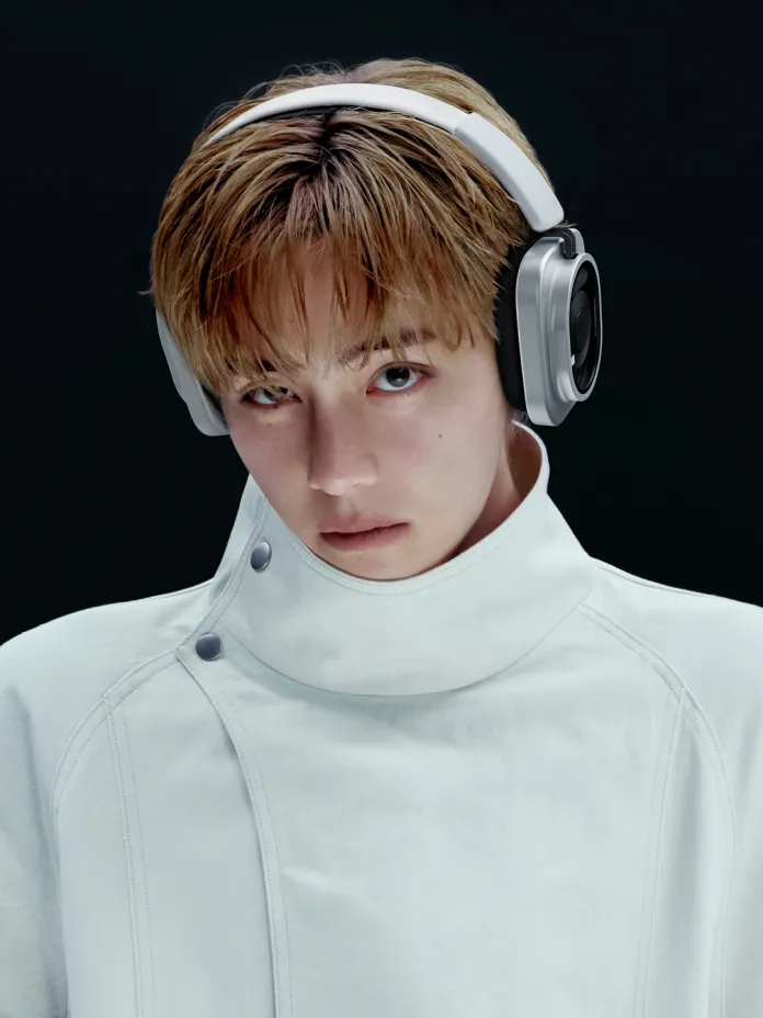 Close-up portrait of BTS V wearing headphones with a serious expression, dressed in a high-collared white jacket against a dark background.