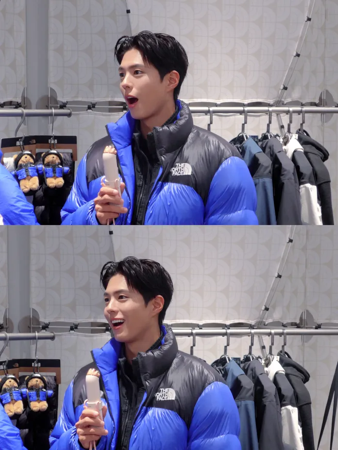 Park Bo-gum with black hair, wearing a blue and black puffy jacket, is smiling excitedly while holding a small object, in a clothing store setting.
