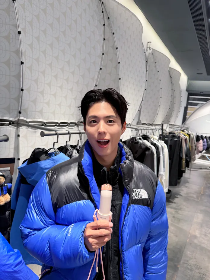 Park Bo-gum wearing a blue and black puffy jacket is smiling and holding a small device resembling a hairdryer, with clothing racks in the background.