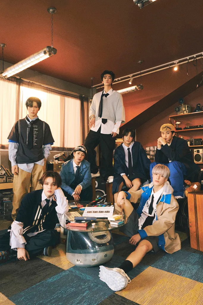 NCT DREAM posing in a cozy, vintage setting filled with shelves of records and retro decor. The individuals are dressed in a mix of casual and stylish outfits, showcasing a blend of modern and nostalgic fashion. They are arranged around a table cluttered with various items, including a vintage radio and colorful stationery.