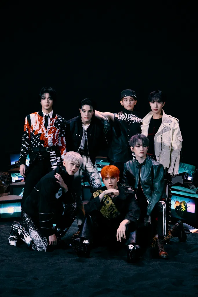 NCT DREAM posing together, dressed in stylish and diverse outfits against a dark background. Some members have colorful hairstyles, and they appear enthusiastic and confident.