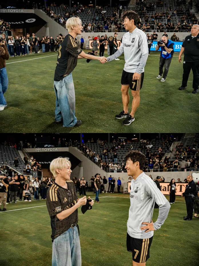 Taemin and Son Heung-min individuals are shaking hands on a soccer field, smiling at each other. Taemin is dressed in a black and gold jersey, while Son Heung-min is in a gray sporting outfit. The stands in the background are filled with spectators.