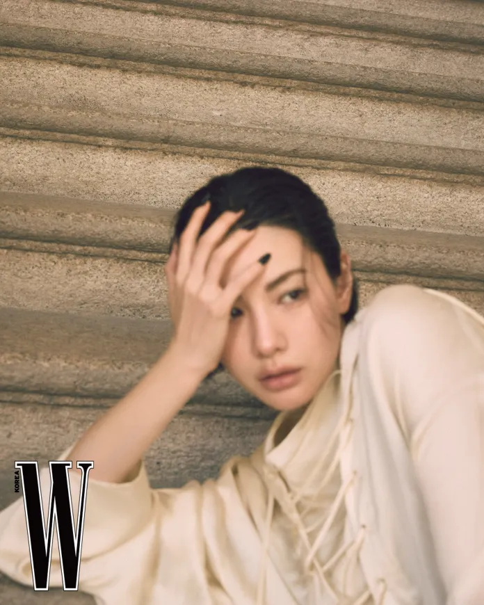 Nana in a light-colored, loose-fitting top sits on stone stairs, leaning against the wall with one hand on her forehead, expressing a contemplative look.