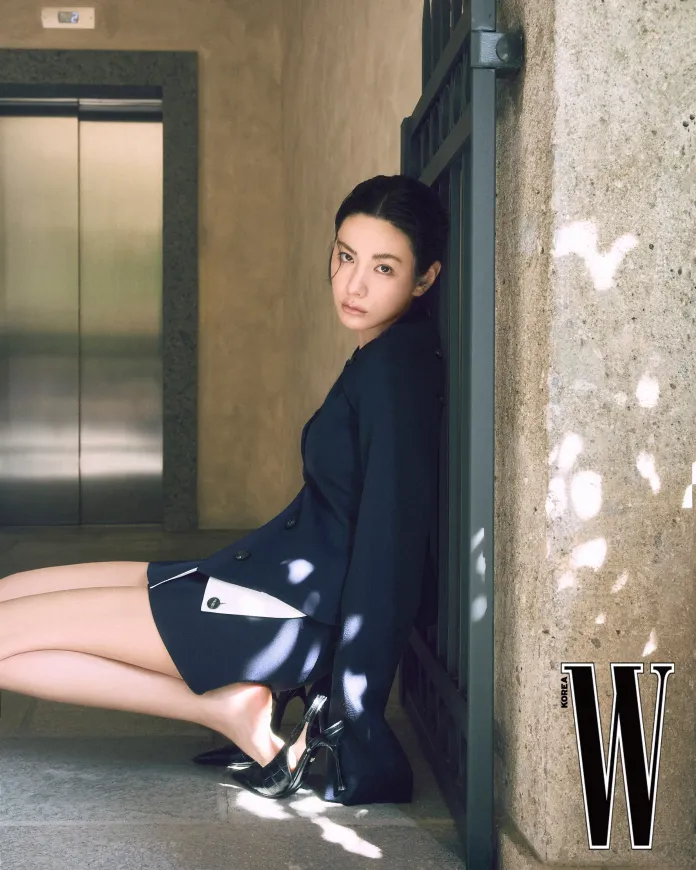 Nana in a navy blazer and skirt sits against a wall, looking at the camera with a thoughtful expression. She is wearing black high heels, and soft natural light creates shadows on the stone surfaces around her.