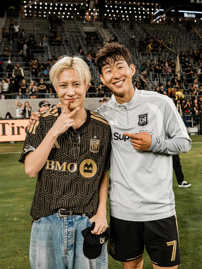Taemin and Son Heung-min posing together on a soccer field, Taemin wearing a black geometric patterned shirt and Son Heung-min in a gray sports jersey. Both are smiling and making gestures with their hands. The background shows a crowd and stadium seating.