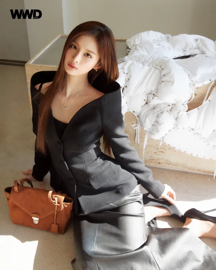 Sullyoon seated on the floor, wearing a black tailored blazer and a fitted black skirt. She has long hair and is holding a brown handbag. The background features soft sunlight and a decorative white fabric piece.