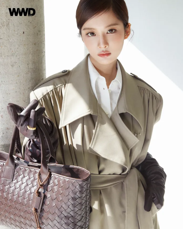 Sullyoon wearing a stylish beige trench coat and leather gloves, holding a brown woven handbag, standing next to a concrete pillar with soft lighting.
