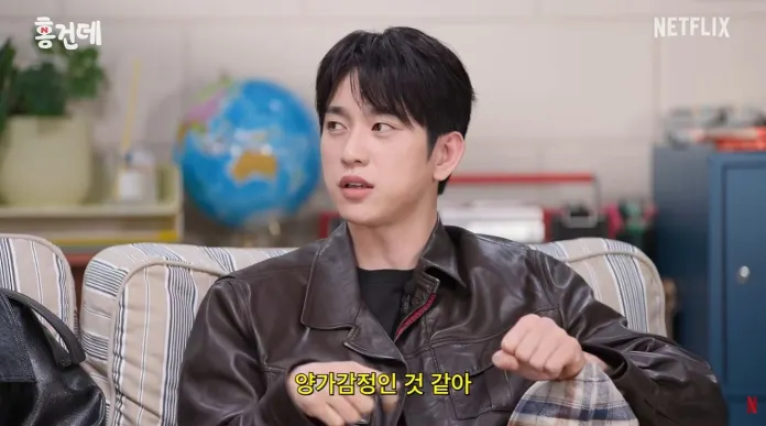 Jinyoung with dark hair and a leather jacket sitting on a couch, looking thoughtfully at something off-camera. A globe is visible in the background.