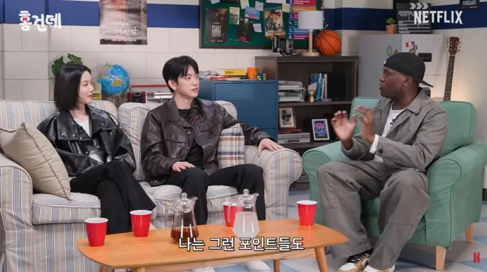 Kim You-jung engaged in a conversation while sitting on a couch. The setting features a cozy living room atmosphere with colorful decor, including a globe and sports items in the background. The individuals are dressed in stylish casual outfits and there are red cups on a table in front of them.