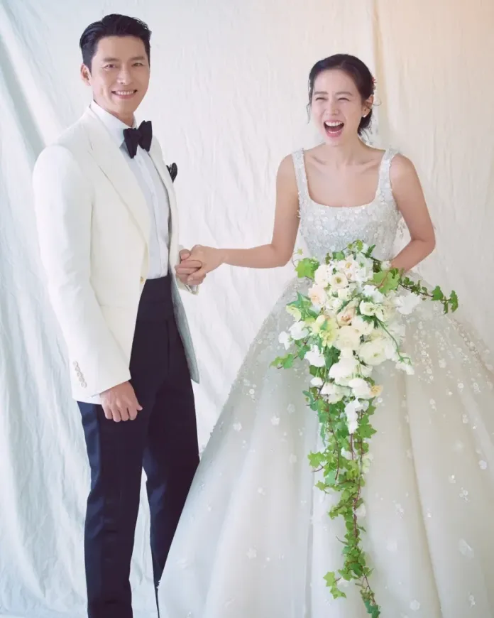 Hyun Bin and Son Ye-jin holding hands, the groom in a white tuxedo and bow tie, and the bride in a sparkling, floral-embellished wedding gown, with a bouquet of white flowers and greenery.