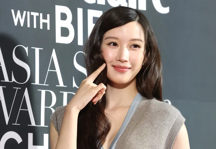 Moon Ga-young with long dark hair is smiling and touching her cheek, standing in front of a dark background with text that likely promotes an event or award show.