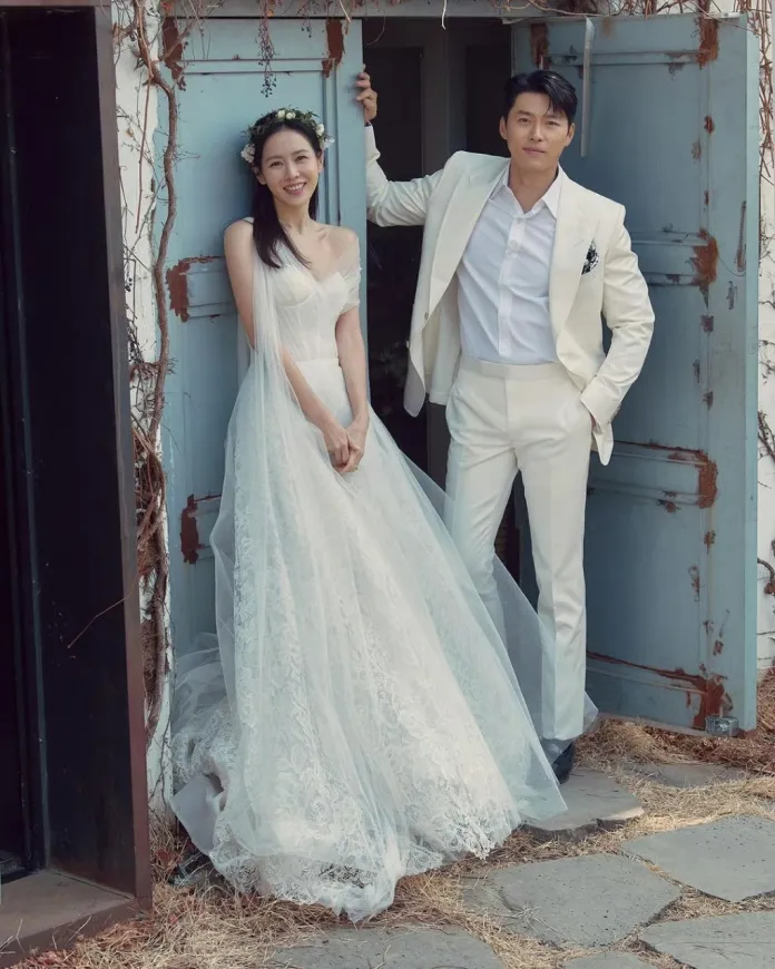 Hyun Bin and Son Ye-jin poses in front of an old door, the bride in a flowing white lace gown with a floral crown, and the groom in a stylish white suit.
