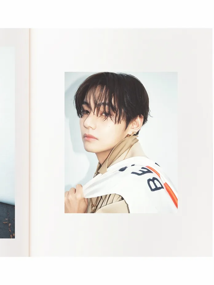 BTS's V with shoulder-length hair, wearing a beige jacket and holding a white garment casually over his shoulder, set against a light background.
