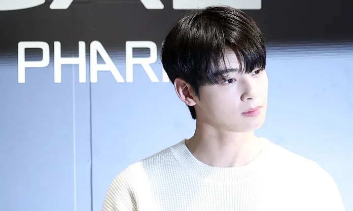 Cha Eun-woo with dark hair wearing a white sweater, looking thoughtfully to the side at a promotional event with a blurred background featuring the word 'PHARMACEUTICAL'.