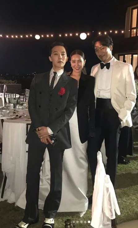 Gdragon his sister and her husband posing together at an outdoor event in the evening. G-Dragon on the left is wearing a gray suit with a red rose, the woman in the middle is dressed in a white gown, and the man on the right is in a tuxedo. Soft lighting and string lights are visible in the background.
