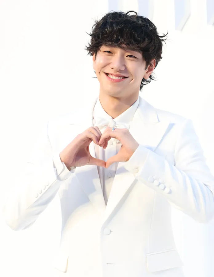 A smiling Nam Yoon-su in a white suit making a heart shape with his hands.