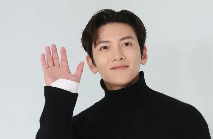 A smiling Ji Chang-wook wearing a black turtleneck sweater, waving with his right hand against a light background.