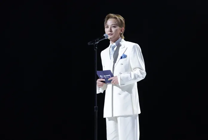 G-Dragon in a white suit and tie stands at a microphone on stage, holding a card and speaking to an audience against a dark background.