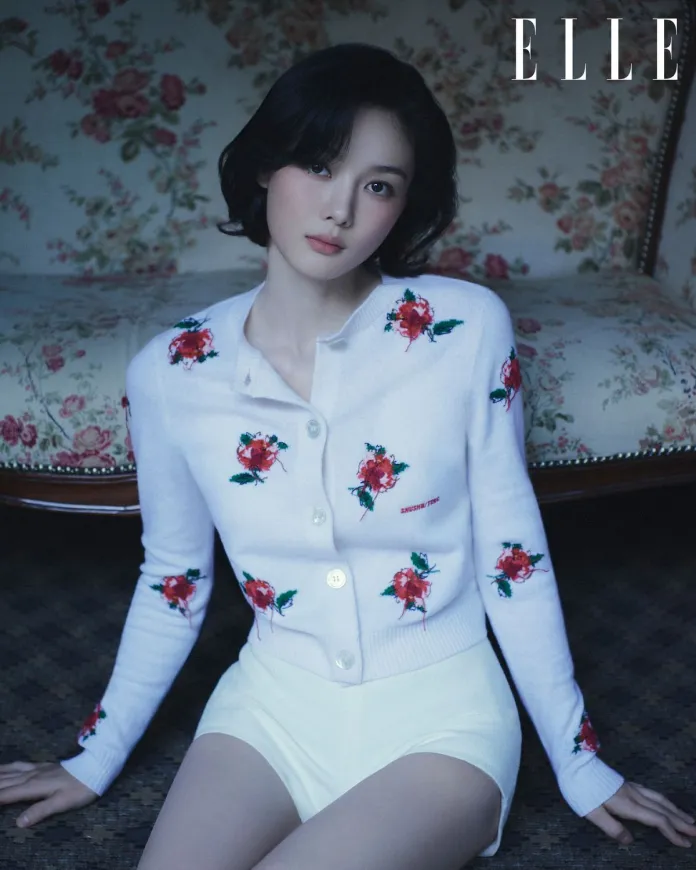 Kim You-jung with short dark hair sits on a floral patterned couch, wearing a white cardigan with red rose embroidery and white shorts, gazing softly at the camera.