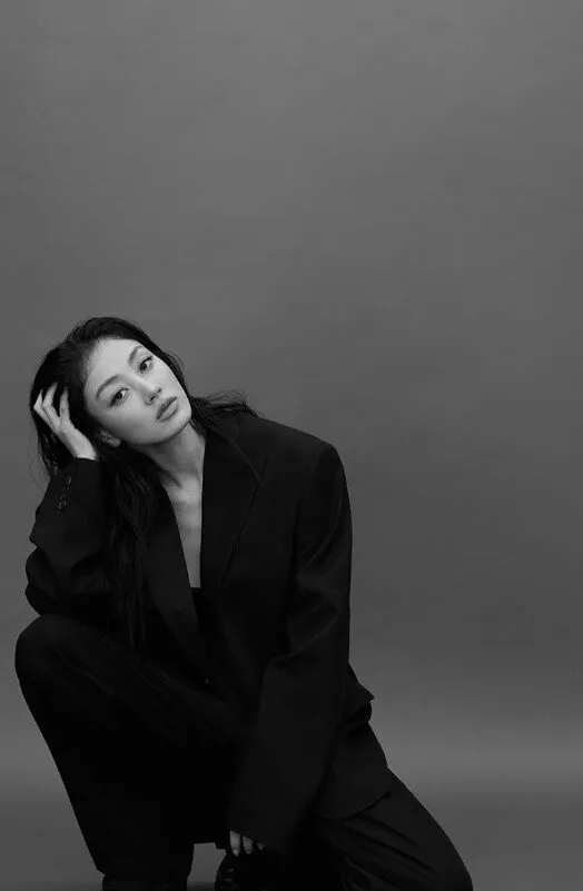 Jihyo in a black oversized suit poses against a plain gray background, with her hand in her hair and a thoughtful expression.