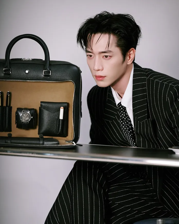 Seo Kang-joon in a pinstripe suit is sitting at a table, looking intently at a stylish black bag displayed on the table. The bag features pockets holding various items, including a watch and writing instruments.