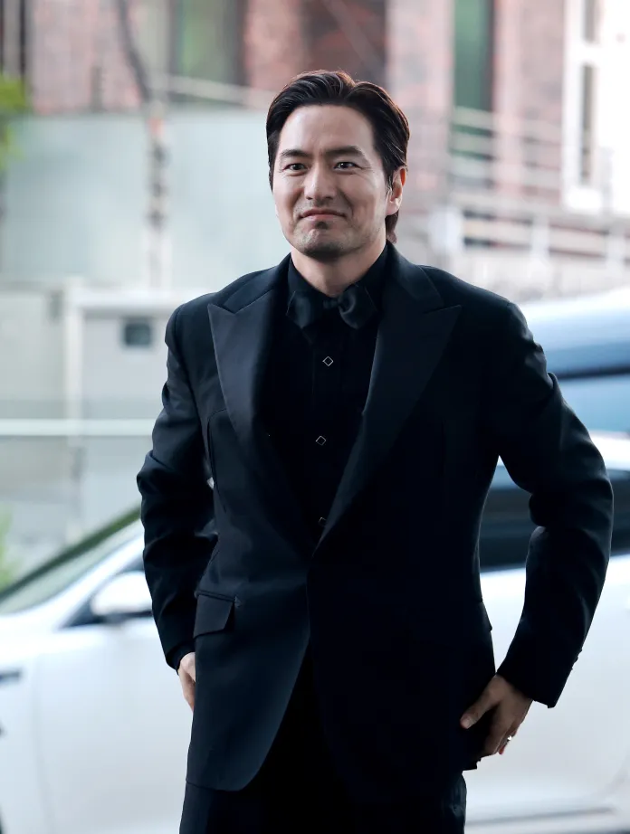 Lee Jin-uk wearing a black tuxedo smiles while standing outdoors, with a car parked in the background.