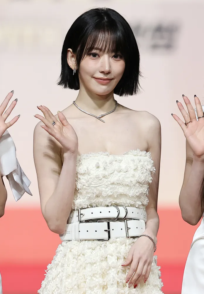 Sakura with short black hair wearing a strapless white dress with a textured design, smiling and waving at the camera. She is accessorized with a necklace and rings.