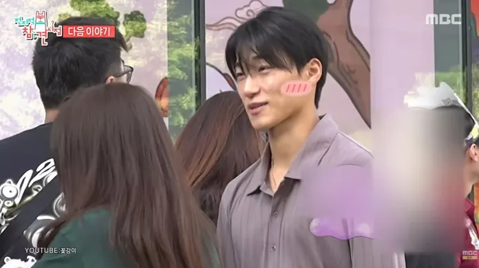 ATEEZ’s San with dark hair, wearing a light gray shirt, smiles while interacting with others in a social setting. Subtle blush marks are illustrated on his cheeks, adding a playful touch to the scene.