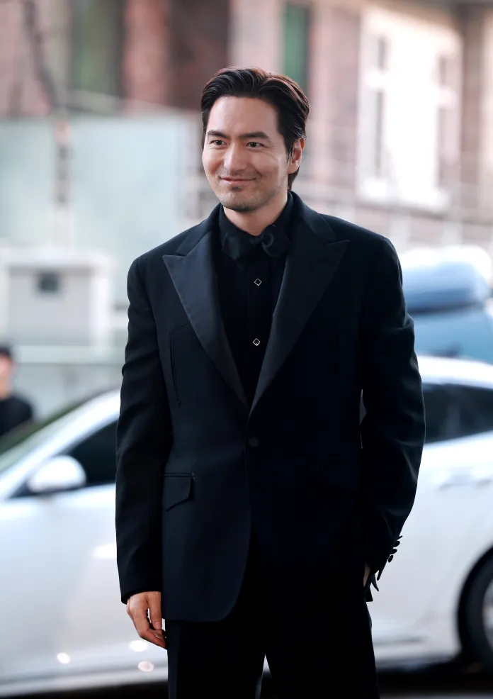 Lee Jin-uk in a black suit smiling in front of a car, with a blurred background of a building.