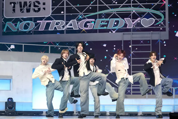 TWS energetically dancing on stage, wearing stylish outfits with loose jeans and coordinated tops, under a colorful backdrop featuring the words 'NO TRAGEDY' illuminated above them.