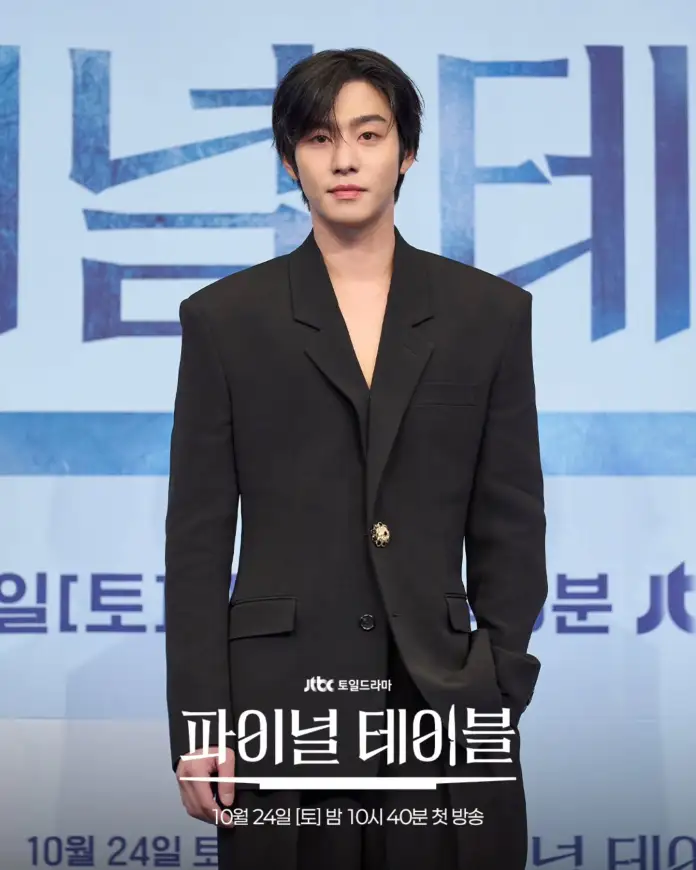 Ahn Hyo-seop with black hair is standing in front of a promotional backdrop for a drama titled 'Final Table'. He is wearing a black oversized blazer and has a neutral expression.