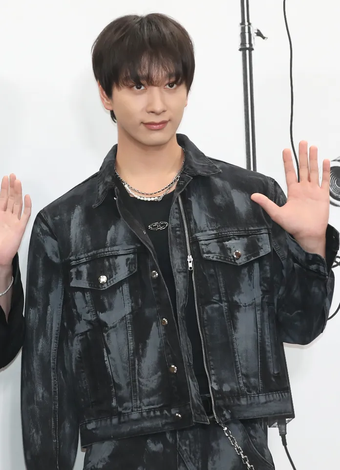 Hwang Chan-sung with black hair wearing a stylish black denim jacket and standing with one hand raised in greeting.