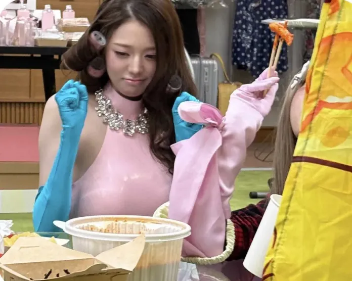 Jang Wonyoung with long brown hair styled in curlers, wearing a pink shiny outfit and blue rubber gloves, is seated at a table covered with food, looking at another individual who holds a piece of food with chopsticks.