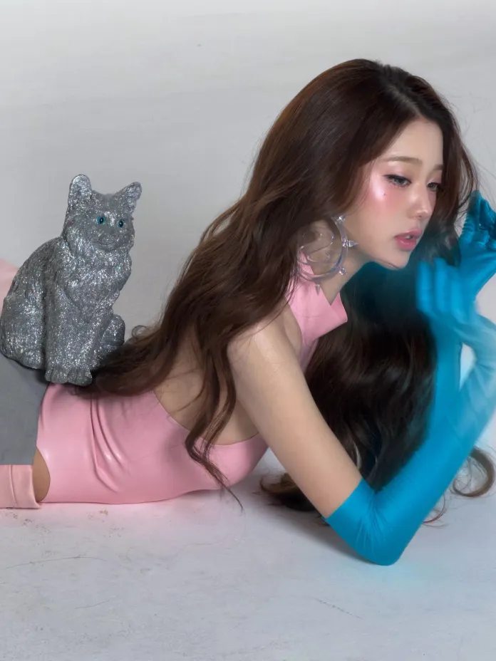 Jang Wonyoung with long hair is lying down, wearing a pink sleeveless top and blue gloves. A shiny gray cat sculpture is positioned on her back.