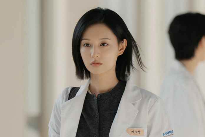 A female doctor wearing a white lab coat and a name tag, with a serious expression, standing in a hospital corridor.