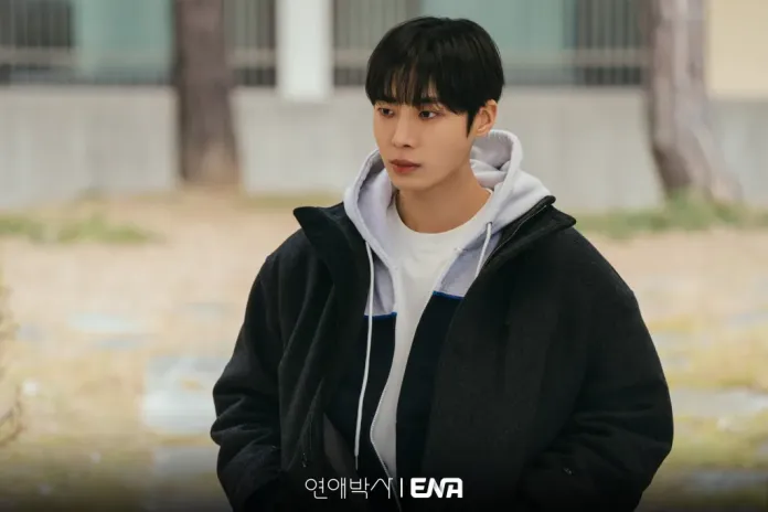 A young man with dark hair wearing a black coat over a gray hoodie and white shirt, standing outdoors with a serious expression.