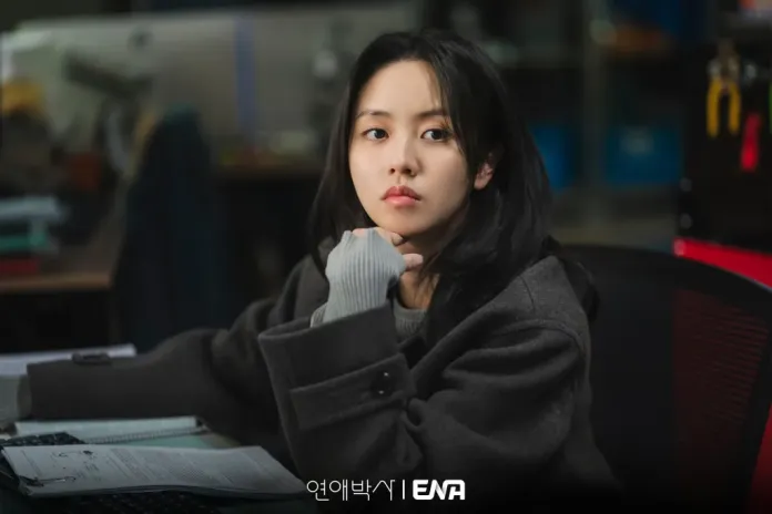 A woman with long black hair sits at a desk in an office, resting her chin on her hand. She is wearing a gray coat and looking thoughtfully at the camera, surrounded by office equipment and papers.