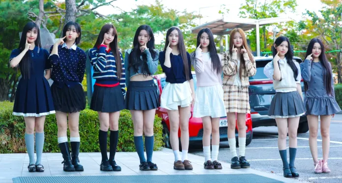 A2O LITE members standing in a row, each making a playful gesture with their fingers to their cheeks. They are dressed in fashionable outfits and skirts, with a variety of colors and styles. The background features greenery and a car.