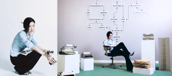 Chaeryeong in a light blue shirt and black pants is sitting on a chair, holding a notepad and looking thoughtful. In the background, there is a wall with a flowchart and several stacks of papers and typewriters around her.