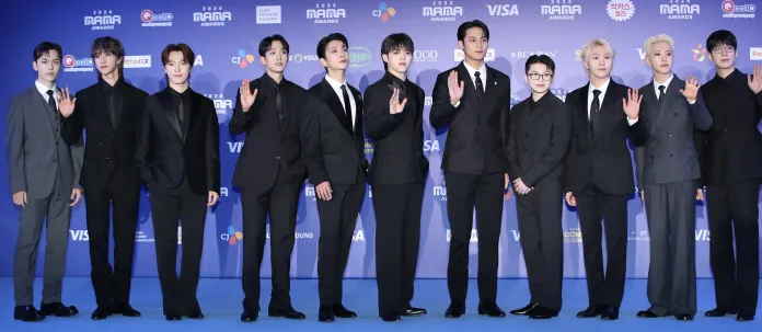 SEVENTEEN members elegantly dressed posing together on a blue carpet at the 2024 MAMA Awards, waving to the camera.