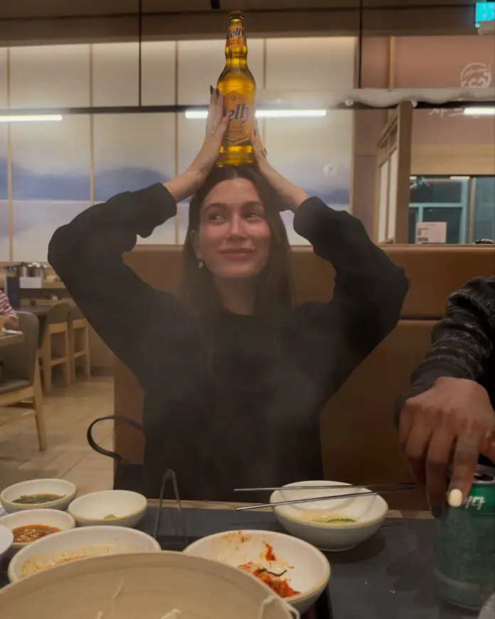 A woman playfully balancing a bottle on her head while sitting at a dining table with various dishes and a steaming pot.