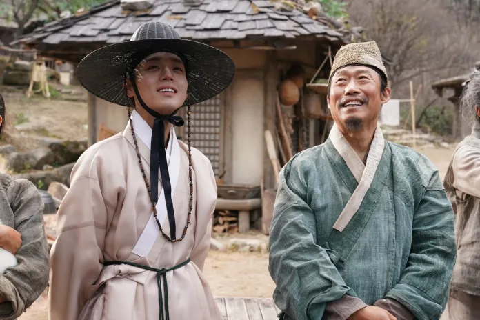 Park Ji-hoon in traditional attire and a wide-brimmed hat smiles joyfully while standing next to an older man in a green robe, both appearing to enjoy a moment together outdoors in a village setting.