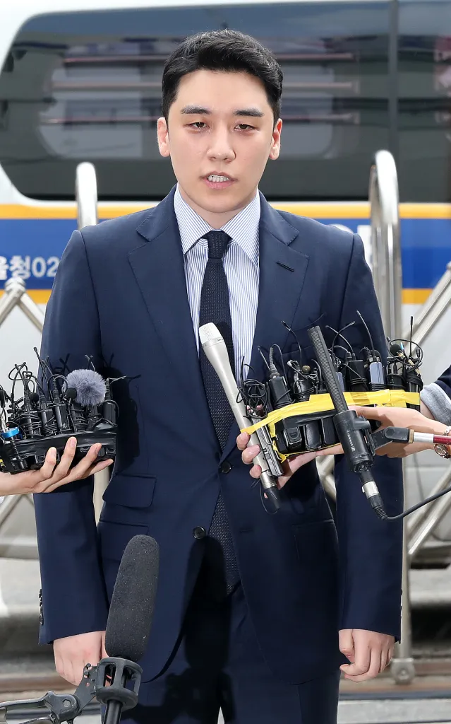  Seungri in a suit stands in front of a group of reporters holding microphones, indicating a press conference or interview. He has a serious expression and is capturing the media attention.