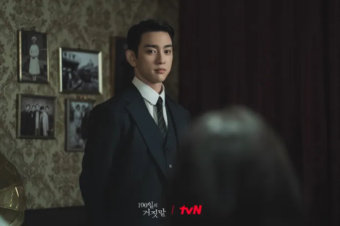 Park Jin-young wearing a formal suit stands in a dimly lit room, facing slightly away from the camera. The background features vintage photographs hanging on the wall, adding to the nostalgic atmosphere.